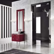 Cuarto de baño con paredes a rayas blancas y negras, lavabo en color blanco sobre modular rojo con cajones. Pared independiente de mármol blanco y bordes de mármol negro.